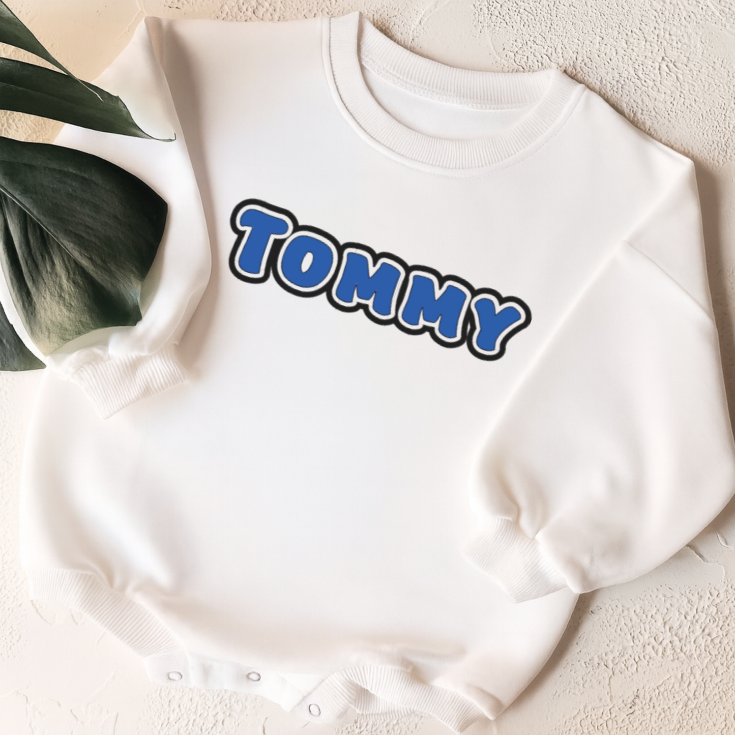 White sweatshirt with 'TOMMY' printed on it, placed on a textured surface with a plant leaf in the corner.