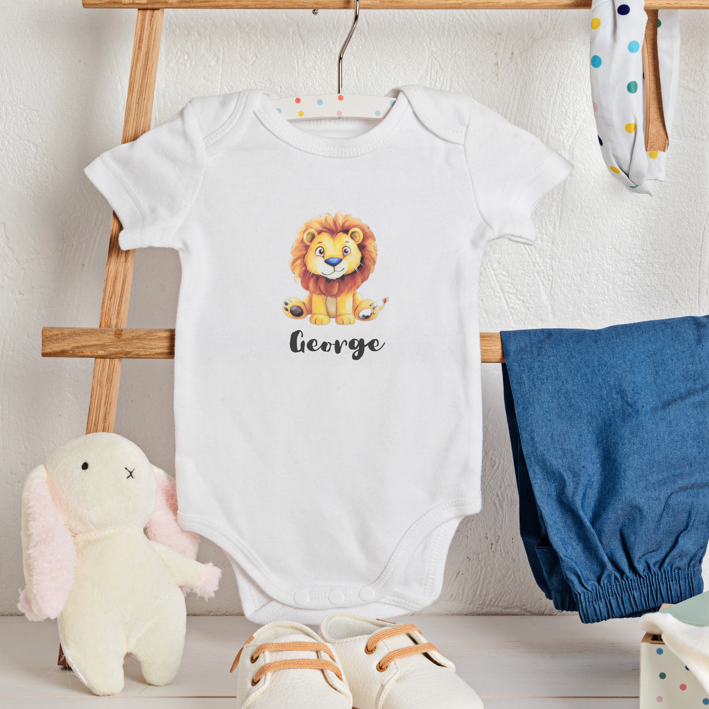 White baby onesie with lion design and 'George' text, blue bloomers, and stuffed animal on a wooden stand.