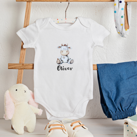 White baby onesie with a cartoon cow design and text, placed on a wooden rack with baby items around.