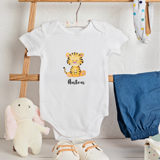 White baby onesie with a tiger design and name, placed on a wooden rack with blue bloomers and a plush toy.