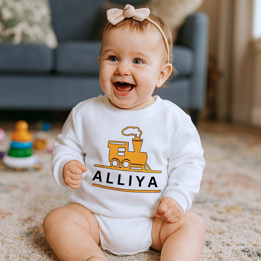 Baby wearing a white onesie with a train design and 'ALLIYA' text, sitting on a carpeted floor.