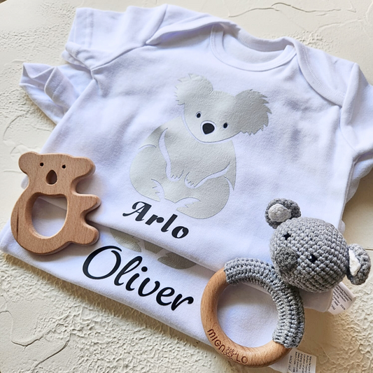 White baby onesie with koala design and 'Arlo Oliver' text, accompanied by wooden and crocheted teething rings.