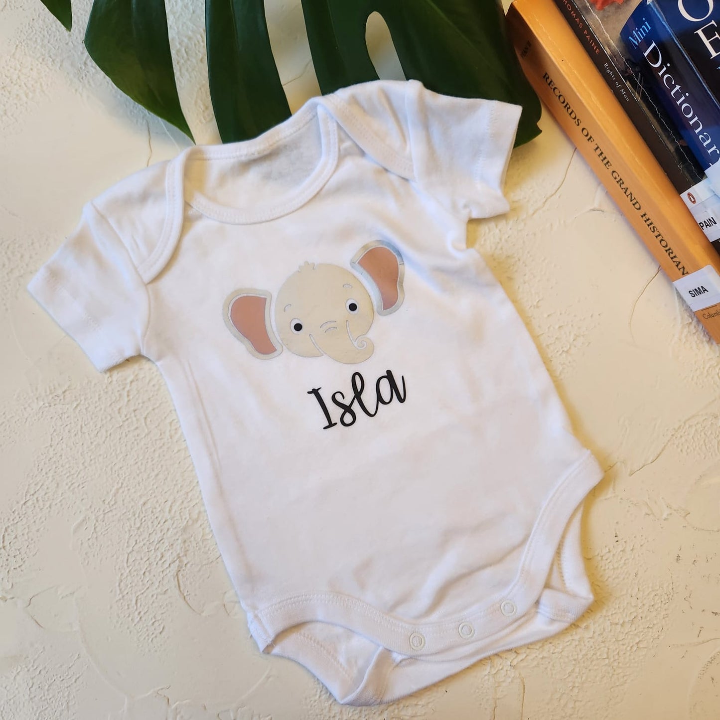White baby onesie with a cartoon elephant design and 'Isla' text on a textured surface.