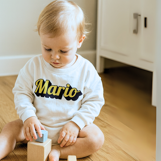 Child wearing a white sweatshirt with 'Mario' printed on it, sitting on a wooden floor.