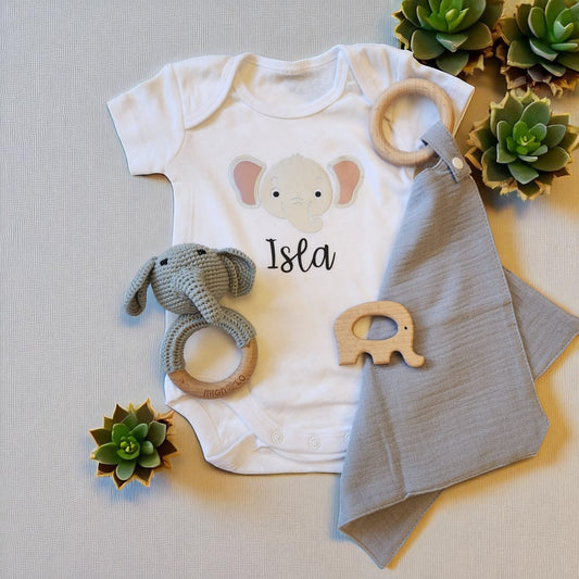 Baby onesie with elephant design, baby blanket, rattle, and wooden toy on a neutral background