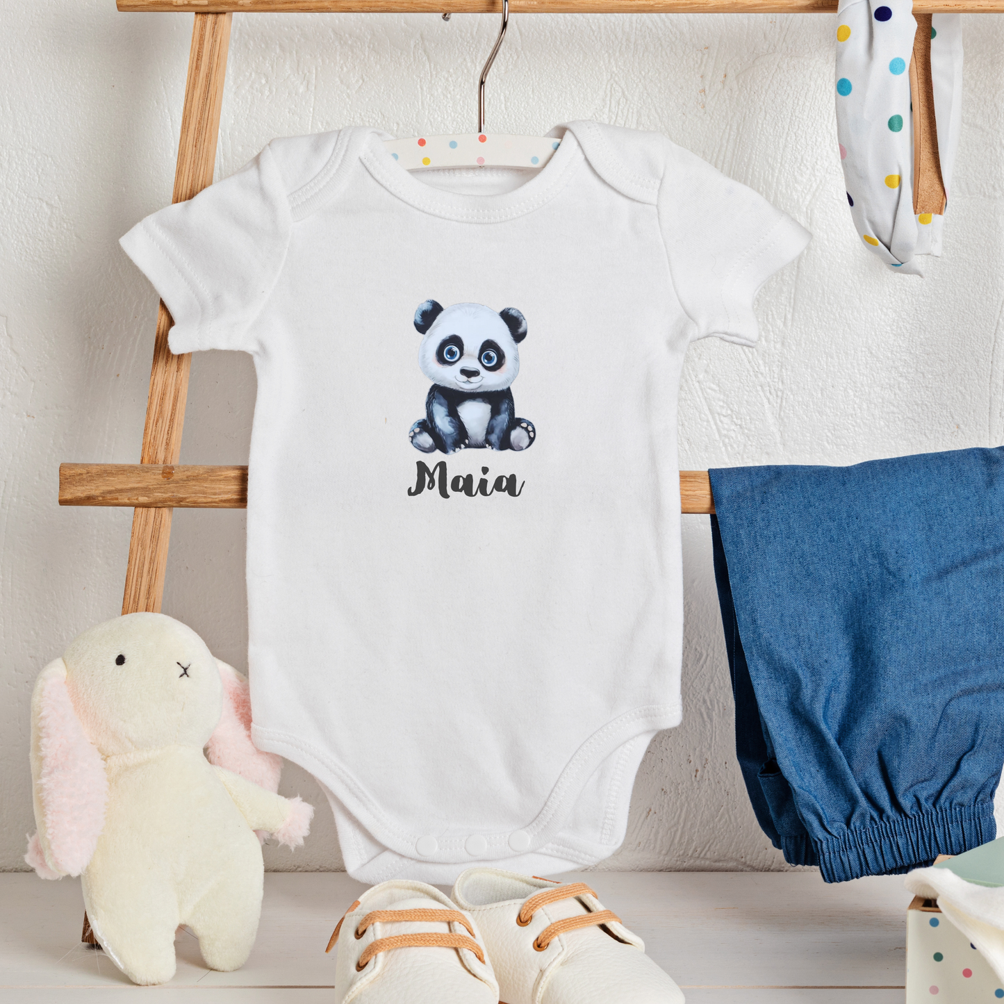 White baby onesie with panda design and 'Maia' text, displayed on a wooden rack with blue bloomers and a plush toy.