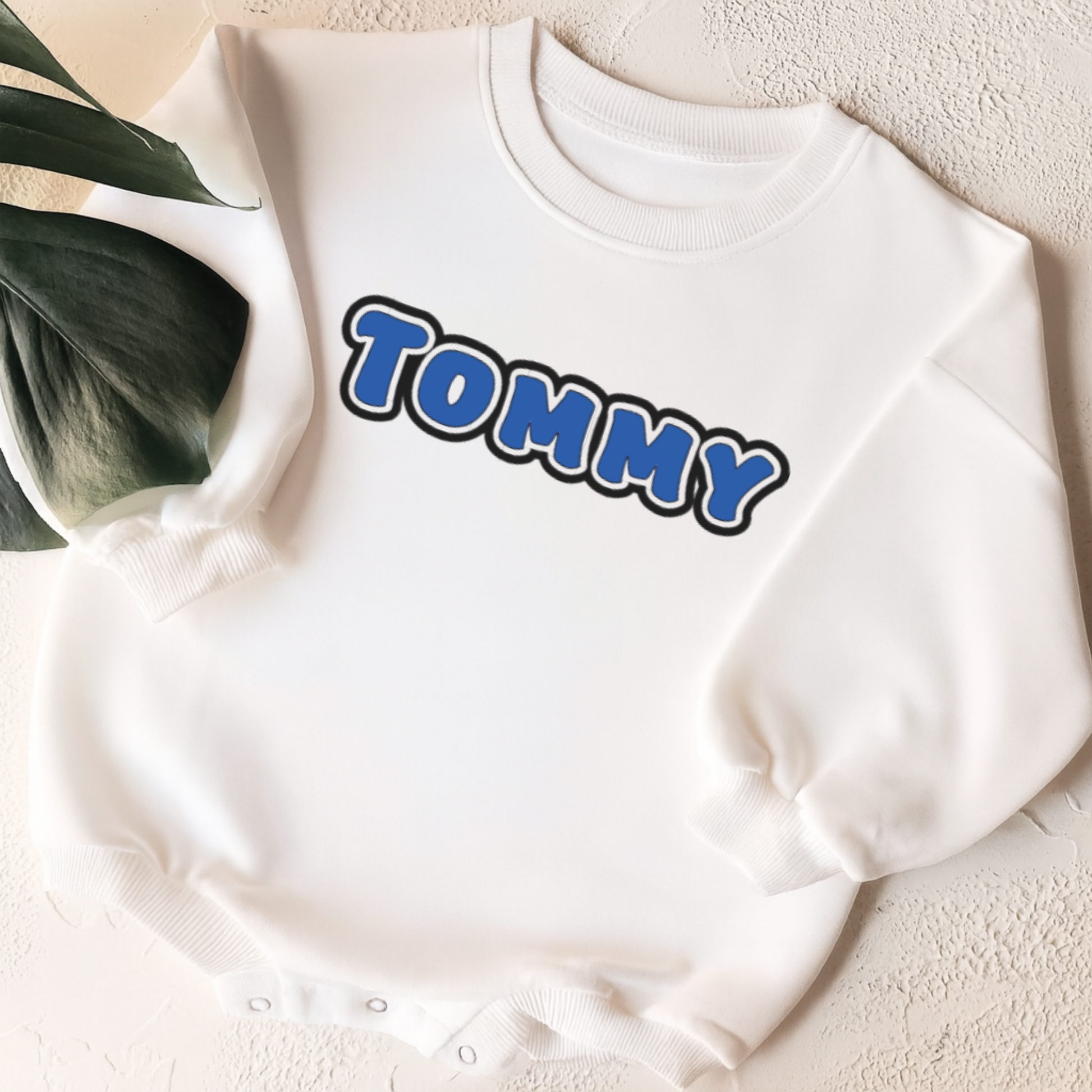 White sweatshirt with 'TOMMY' printed on it, placed on a textured surface with a plant leaf in the corner.