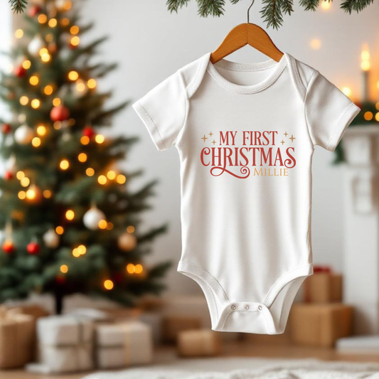 Baby onesie with 'My First Christmas Millie' text on a hanger in front of a decorated Christmas tree.