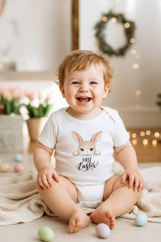 Baby wearing a 'My First Easter' onesie sitting on a blanket with Easter eggs.