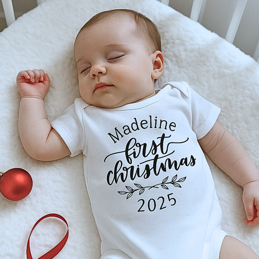 Baby wearing a 'Madeline's First Christmas 2025' onesie on a white blanket with a red ornament.